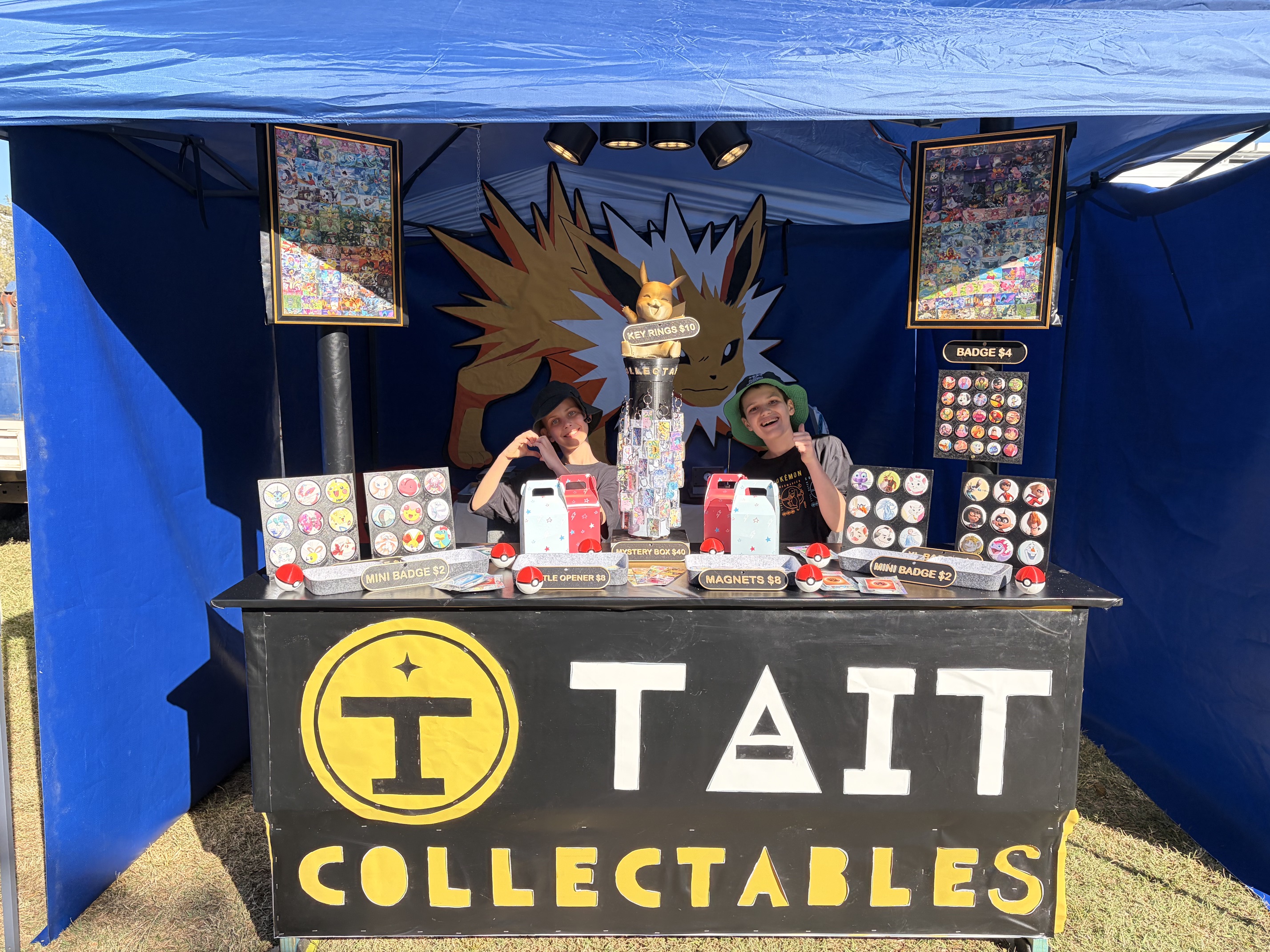 The Tait brothers at their market stall in Bulimba — blue gazebo, Tait Collectables counter, badges, magnets and keychains on the table